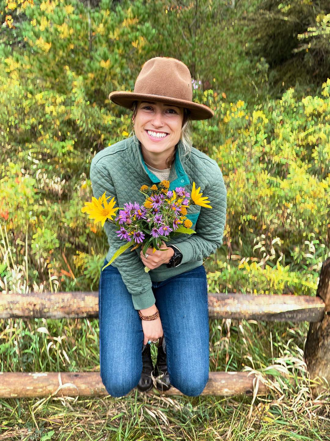 Wild & Well's founder kneeling on a wooden fence in a summer field, holding a bouquet of freshly picked wildflowers