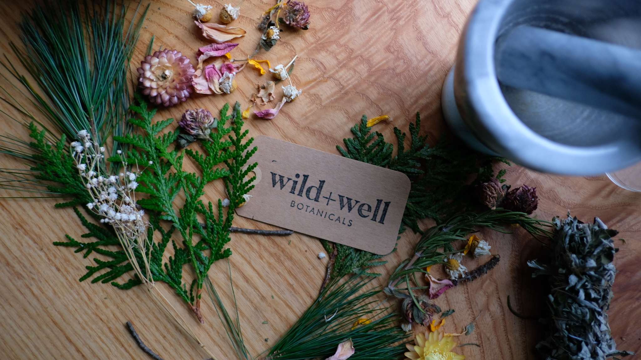 Kraft paper tag surrounded by cedar sprigs, pine, dried flowers and a wooden mortar and pestle, photographed from above