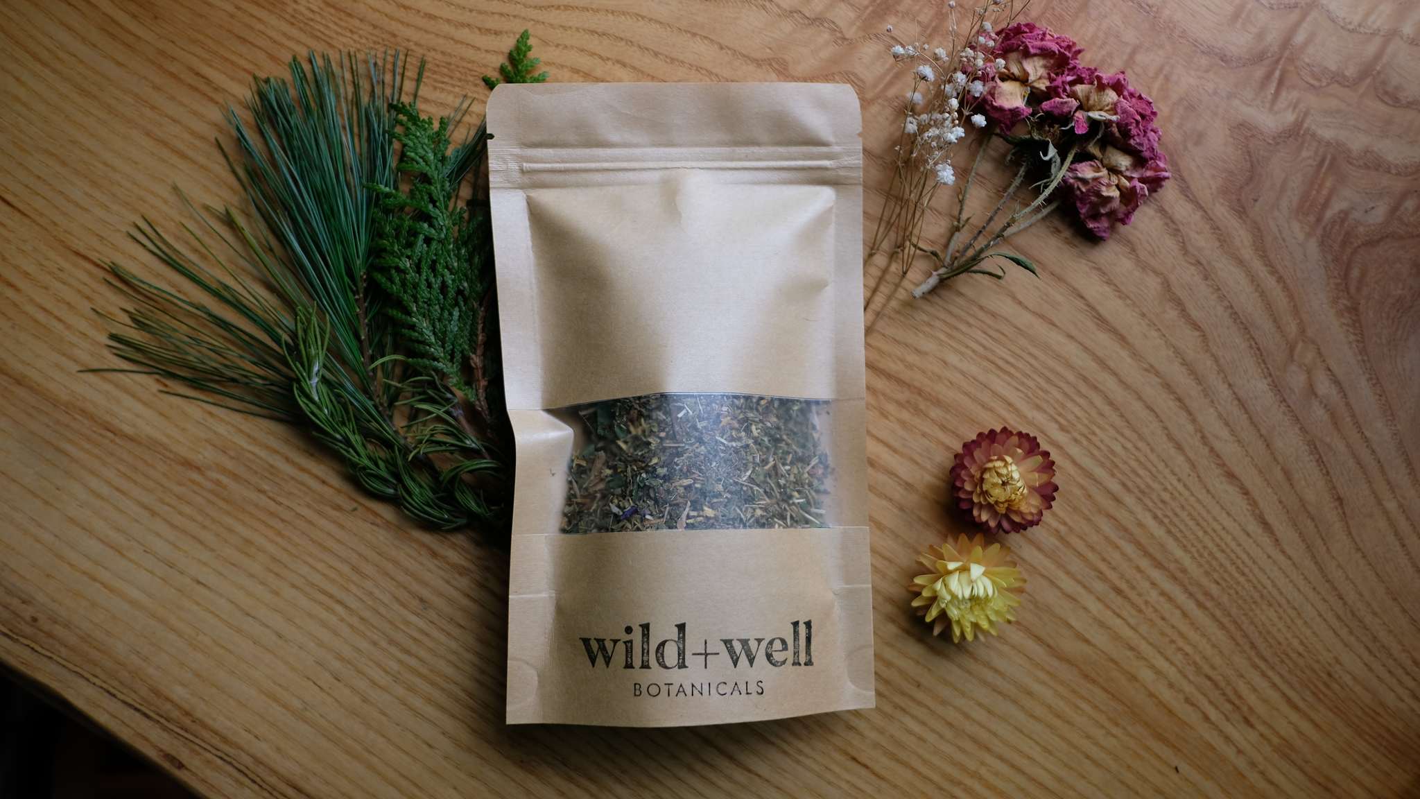 Kraft tea pouch centred on an oak table between a pine bough and a spray of dried flowers